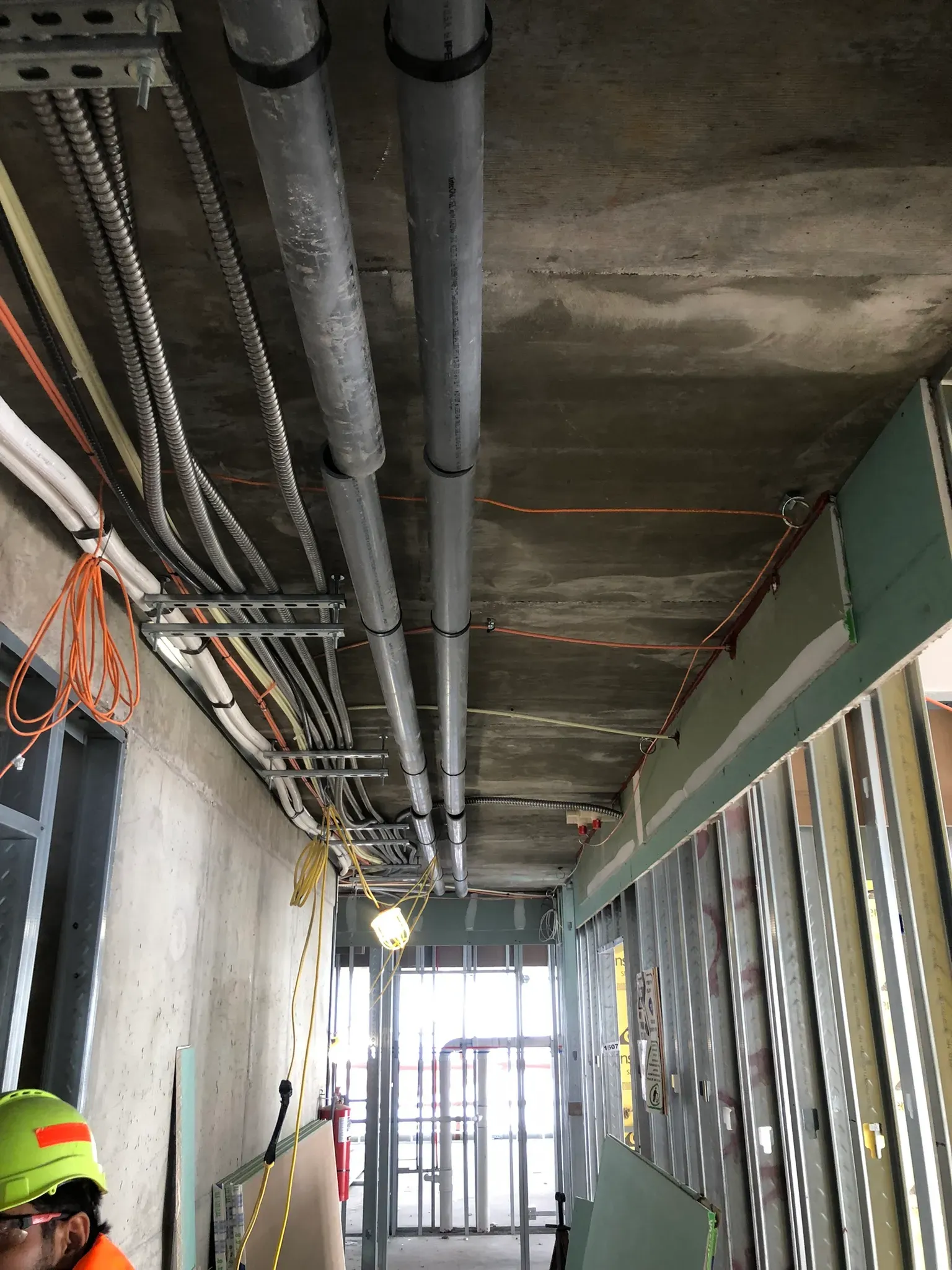 Ceiling view of a commercial construction corridor showing exposed conduit, piping, cable trays, and metal stud wall framing during rough-in