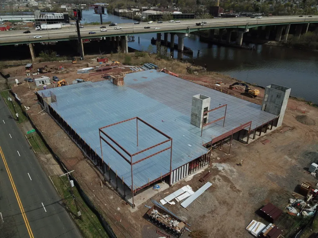 Aerial view of a low-rise commercial building under construction with metal decking, concrete elevator cores, and exposed steel framing beside a river.