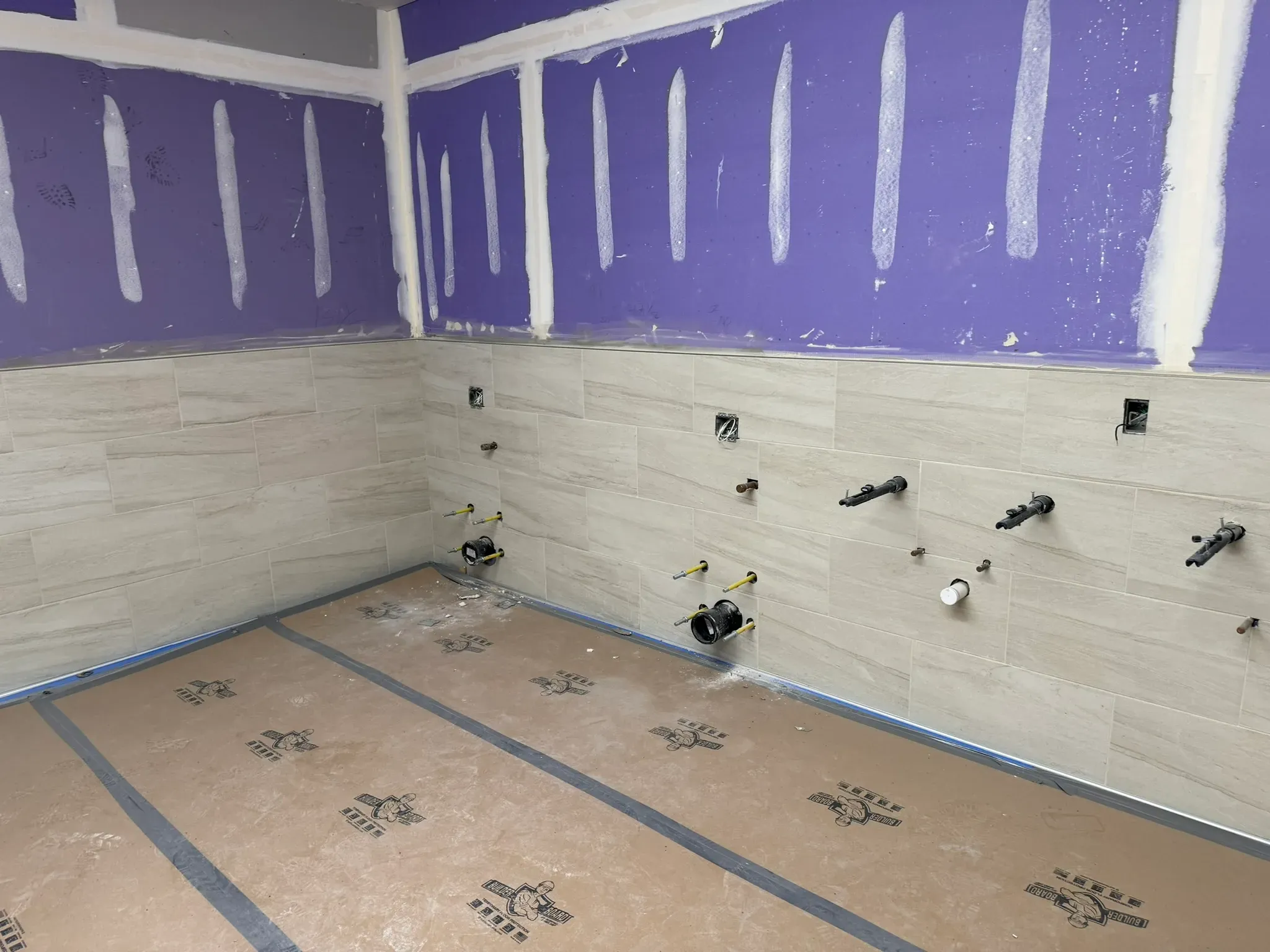 Renovation in progress in a tiled bathroom or utility room, with beige wall tile installed halfway up, exposed plumbing rough-ins and electrical boxes, purple waterproof backer boards above, and floor protection covering the unfinished floor.