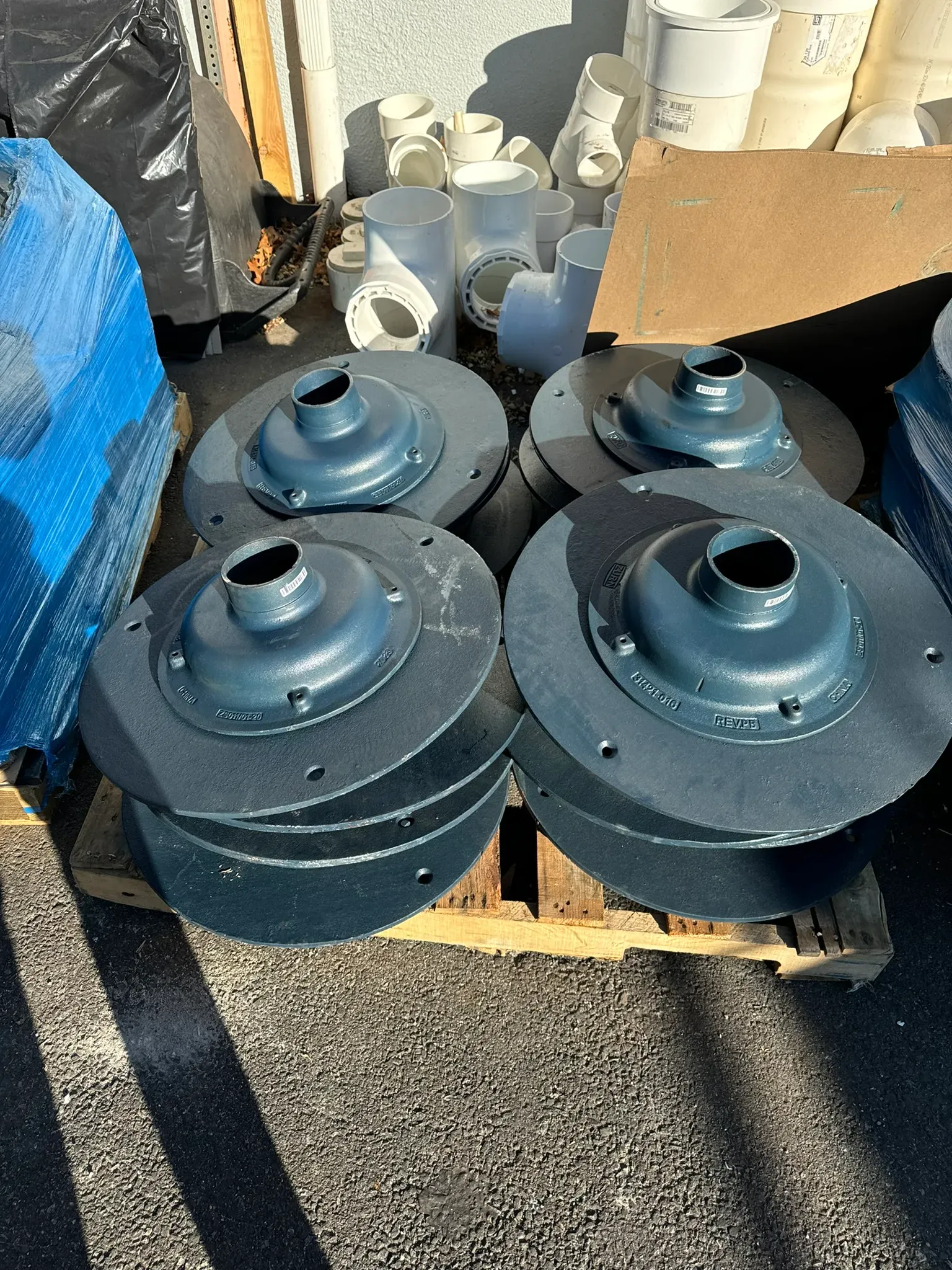 Stack of round metal roof drain components on a pallet beside white PVC plumbing fittings and construction materials.